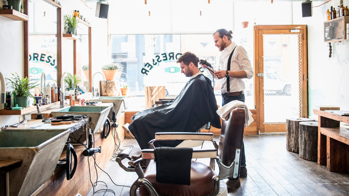 A barber shaves a customer's neck in a barbershop with large windows. The customer wears a dark cape over his clothes. Both people are shown in profile. An empty barber chair is in the foreground.