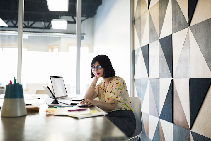 A person sits at a desk and looks at a tablet and a laptop.