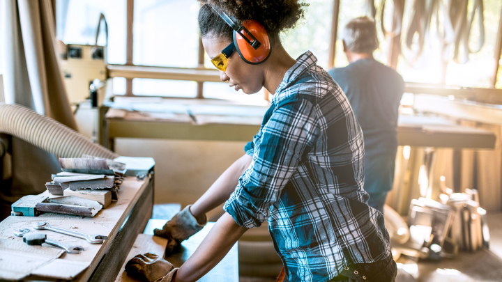 A worker wearing a plaid shirt and safety goggles and ear protection works with wood.