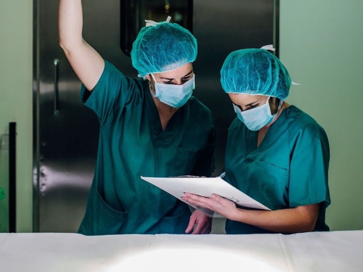 Two health care professionals review medical information to align surgical operating room lights. They are dressed in green surgical scrubs with head covers and face masks.