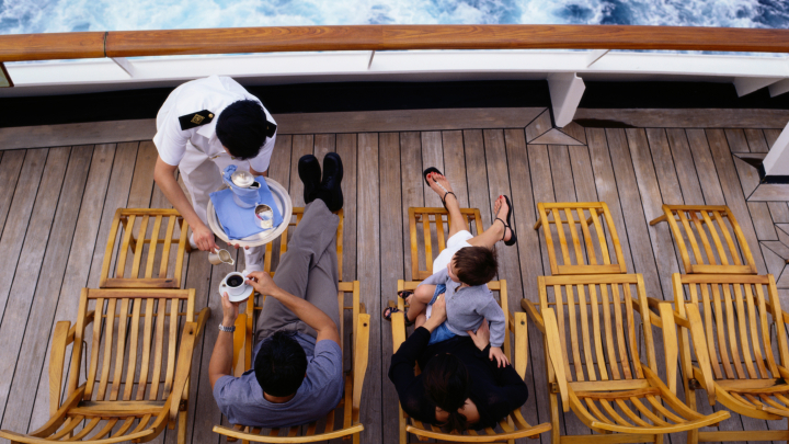 The image shows parents and a child sitting on loungers on the deck of a cruise ship. A server is pouring coffee for the family.