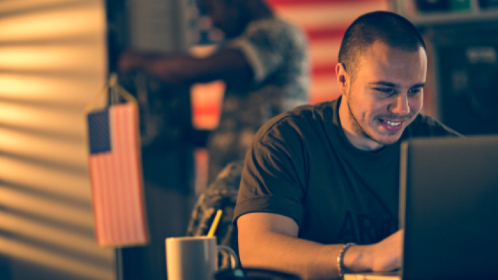The image shows an army soldier in an army t-shirt smiling at something on his laptop. Out of focus in the background, a soldier in uniform is behind him, holding a flag.