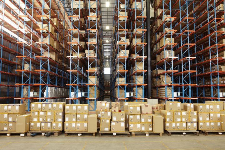 The fully stocked rows of a large warehouse are pictured with stacks of boxes on crates in front of each row.