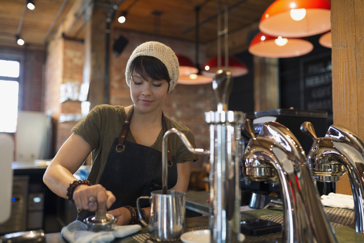 A person muddles ingredients in a coffee shop or small restaurant setting. A milk steamer and two other pieces of equipment are in the foreground.