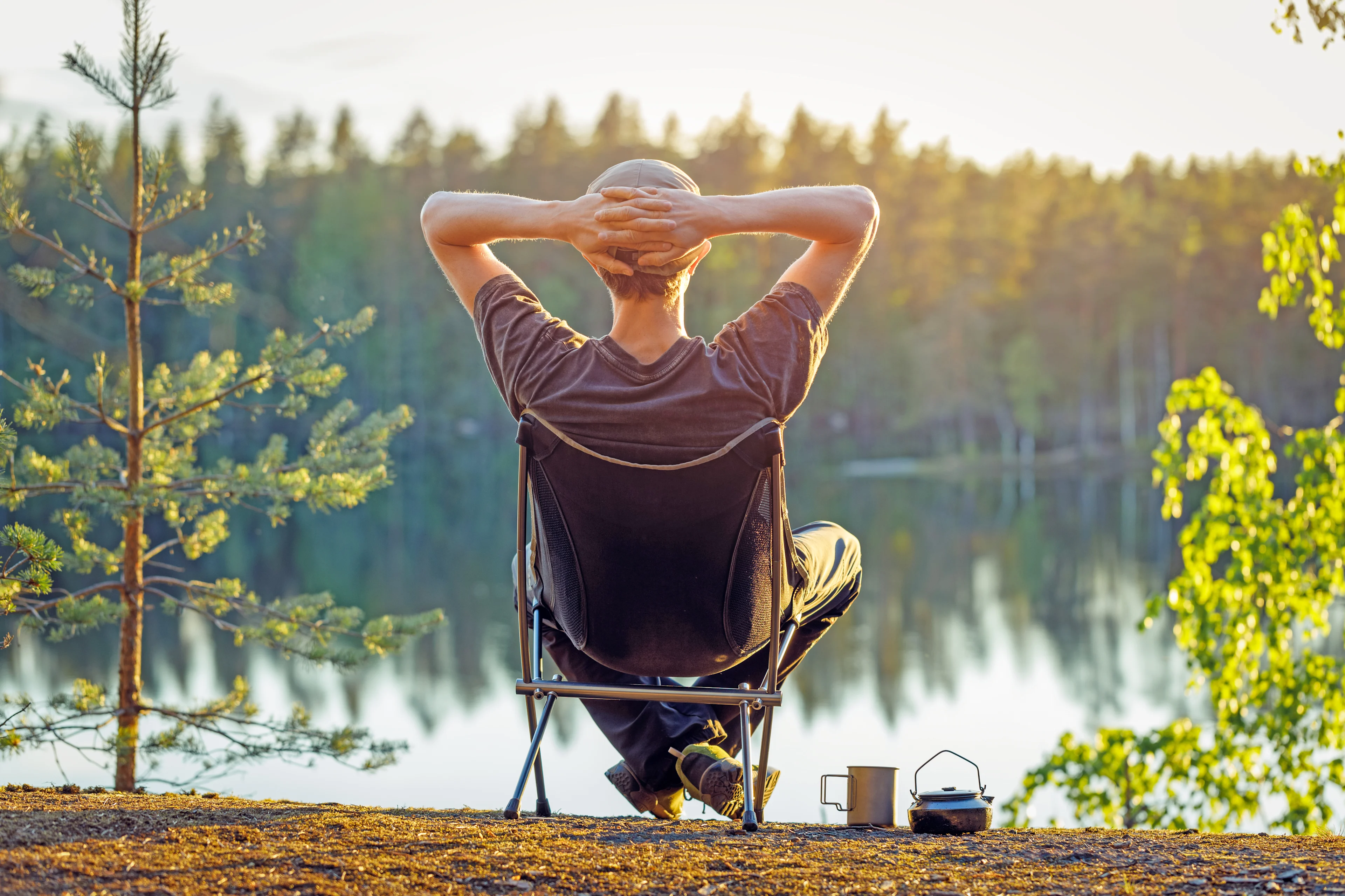 Henkilö istuu retkituolissa järven rannalla auringonlaskun aikaan, kädet pään takana rentoutuneessa asennossa, edessään kahvimuki ja nokipannu.