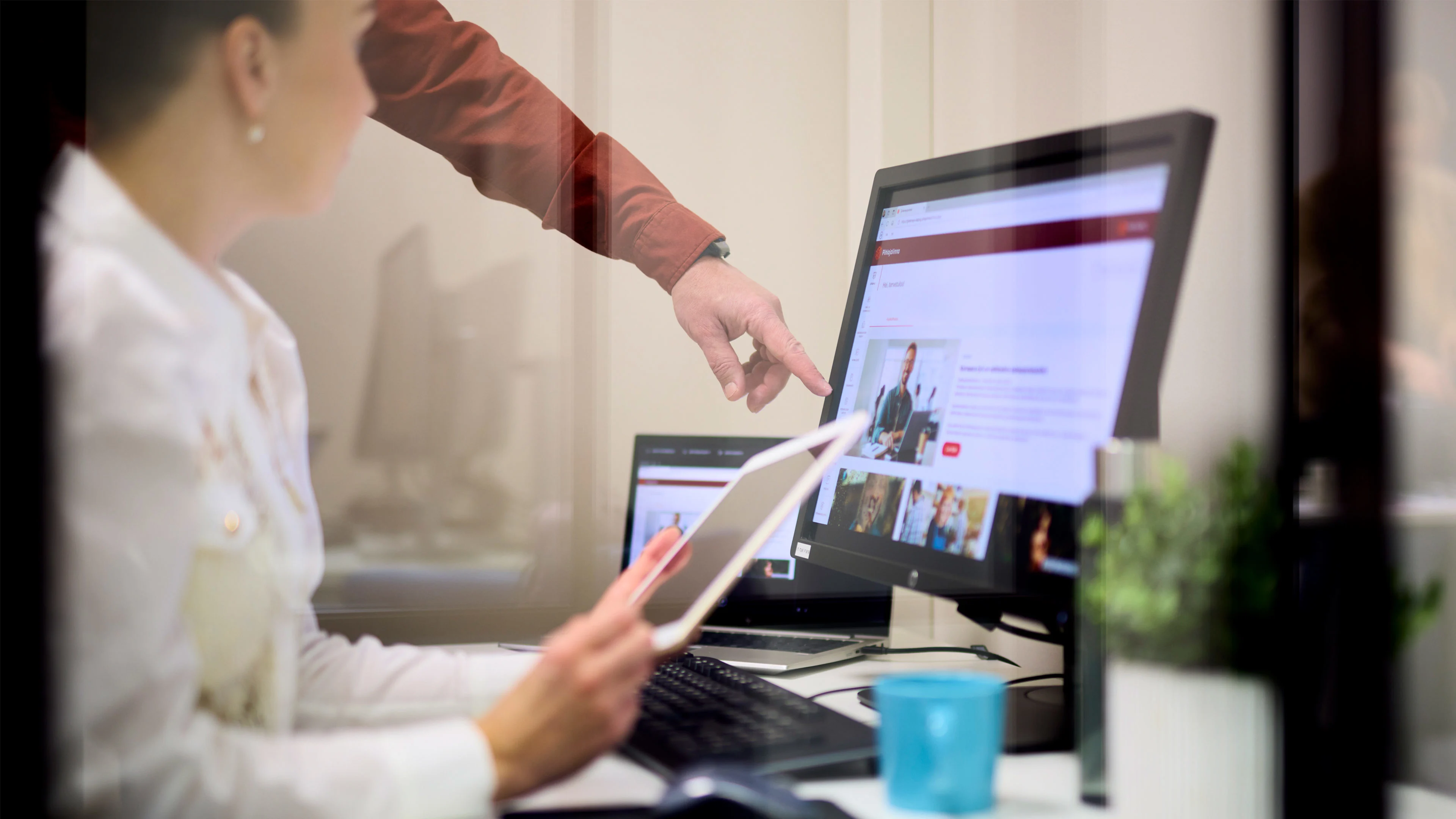 Kaksi henkilöä työskentelee tietokoneella toimistossa, toinen osoittaa näytölle ja toinen pitää tablettia.