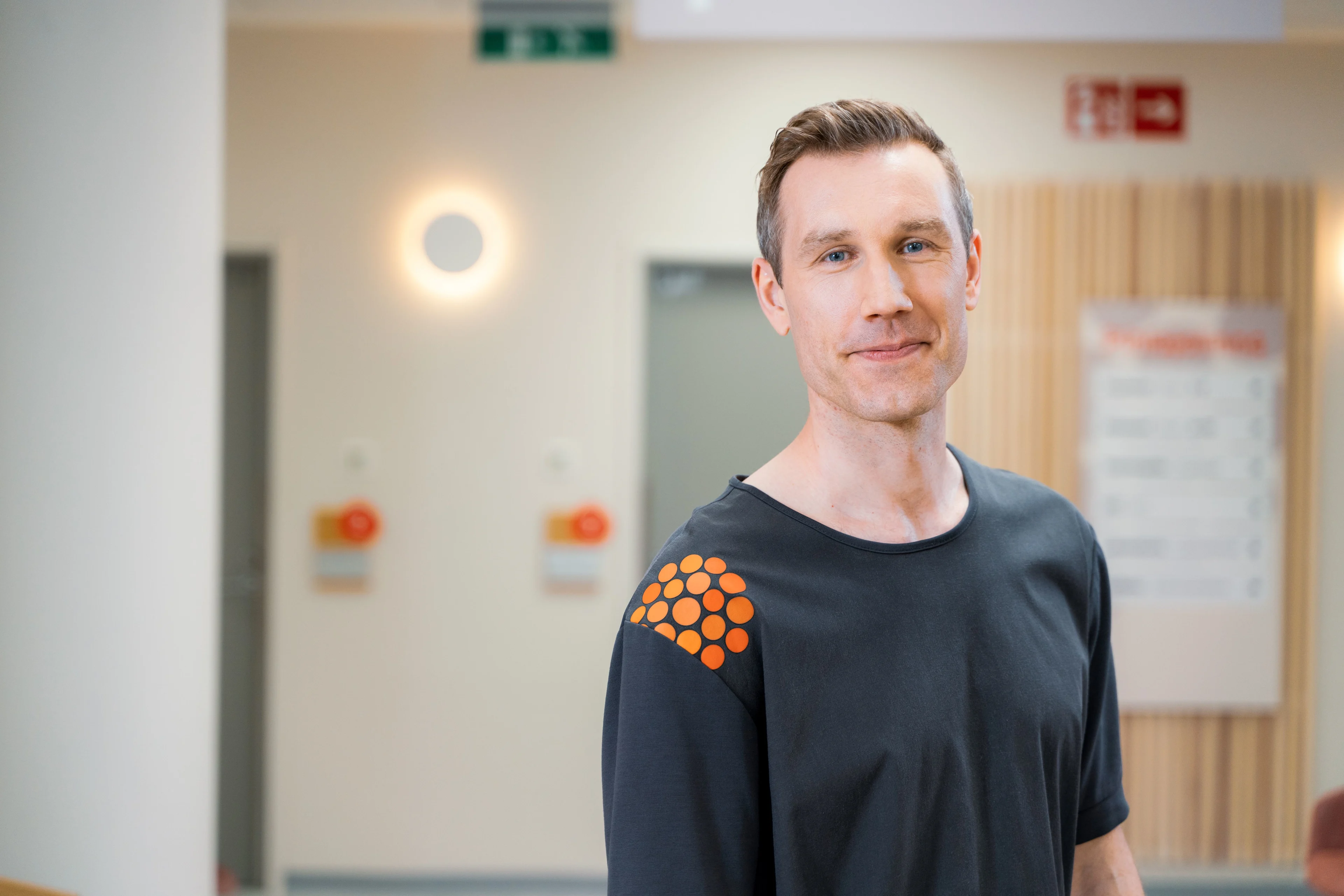 Miespuolinen fysioterapeutti katsoo kameraan tummassa T-paidassa, jossa on Pihlajalinnan logo olkapäässä.