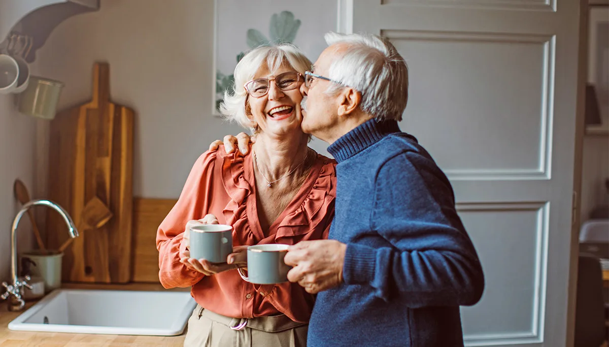 Kaksi vanhempaa ihmistä halaa keittiössä teekupit kädessä.