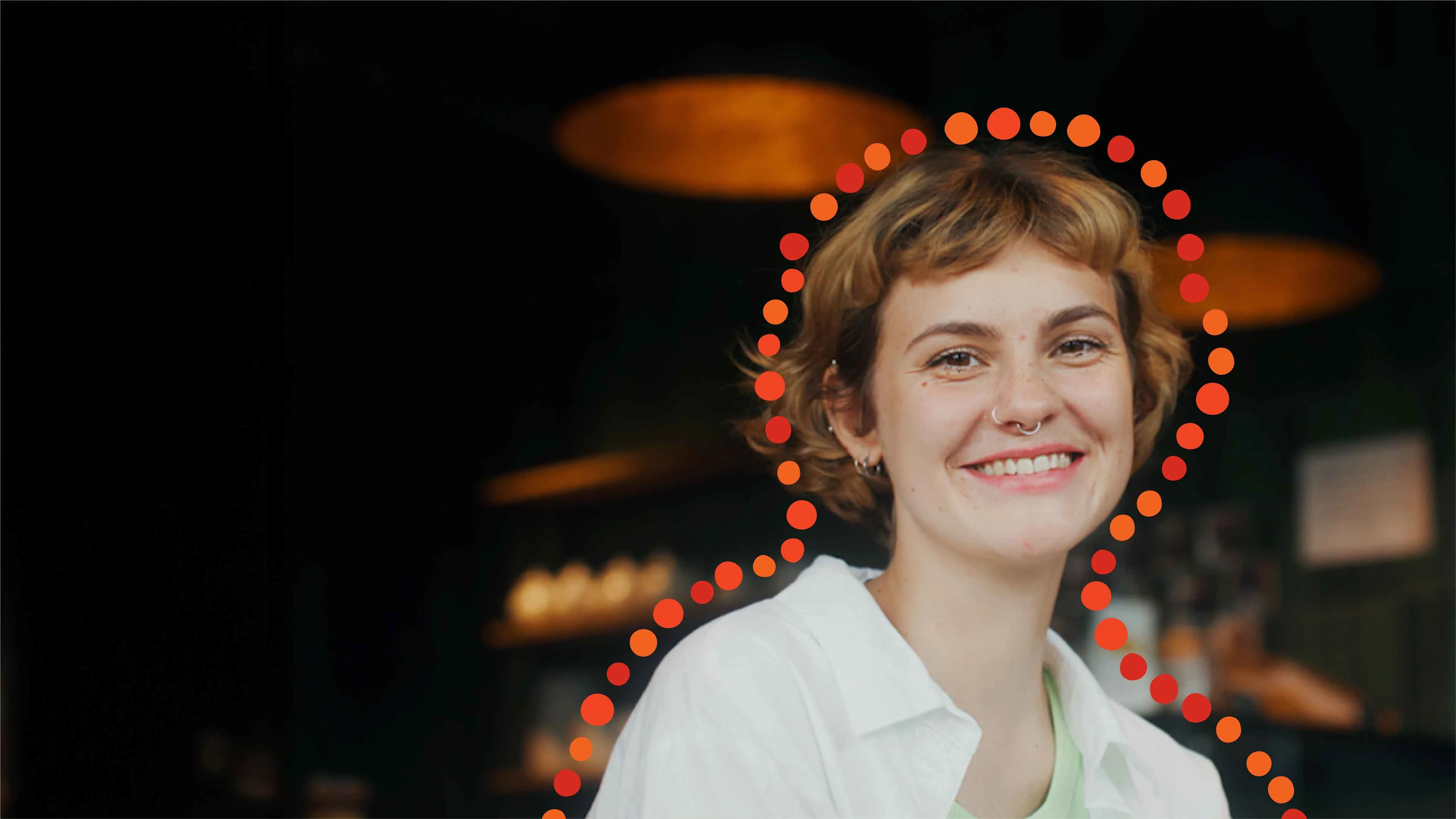 Lyhythiuksinen nainen katsoo hymyillen kohti tummaa taustaa vasten. Naisen ympärillä oranssia pallomaista nauhaa