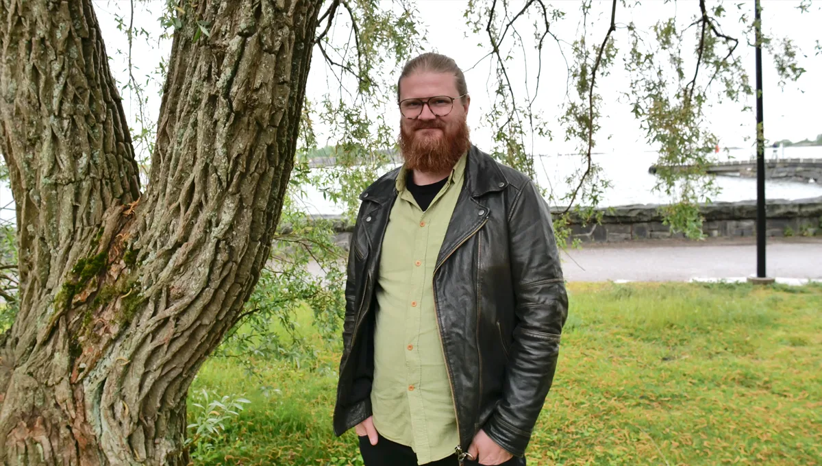 Mies seisoo puun vieressä puistossa, taustalla näkyy merta ja rantakiveystä.