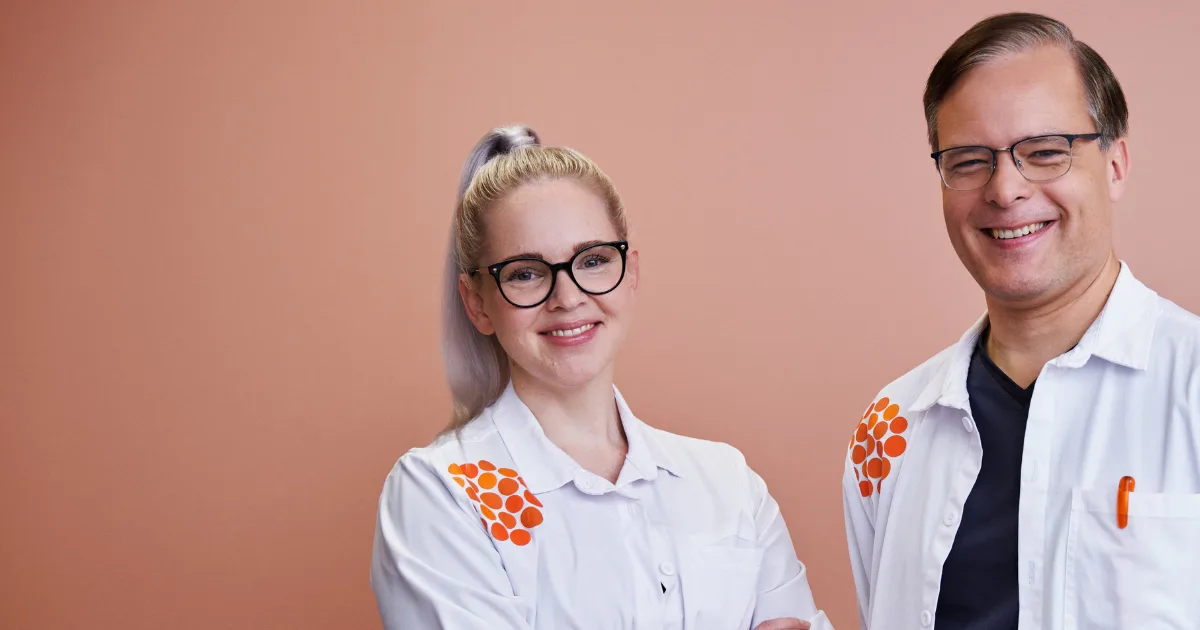 Kaksi hymyilevää terveydenhuollon ammattilaista valkoisissa työasuissaan, vaaleanpunaisen seinän edessä.