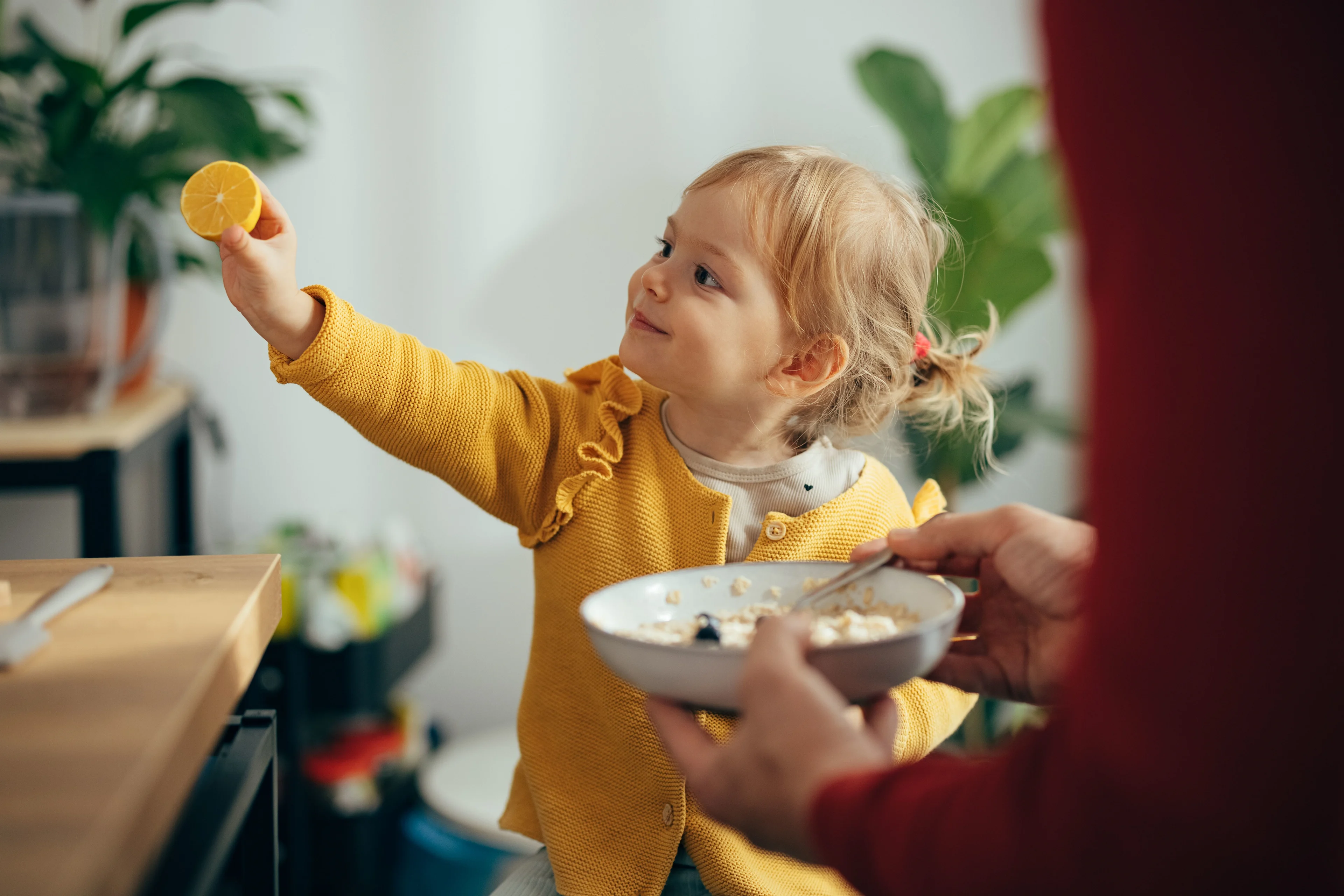 Pieni lapsi pitää sitruunaa kädessään ja hymyilee, samalla kun saa aamiaista kulhosta.