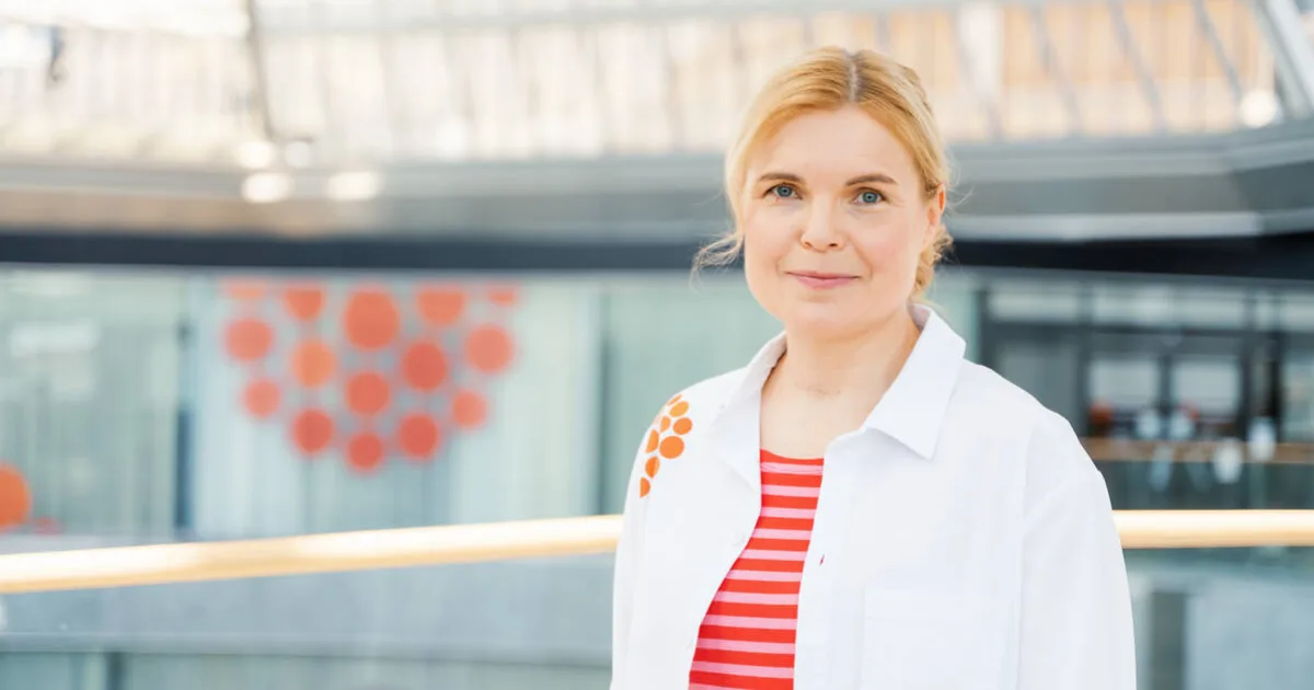 Hymyilevä lääkäri seisoo modernissa sisätilassa, yllään valkoinen takki ja punavalkoinen raitapaita.