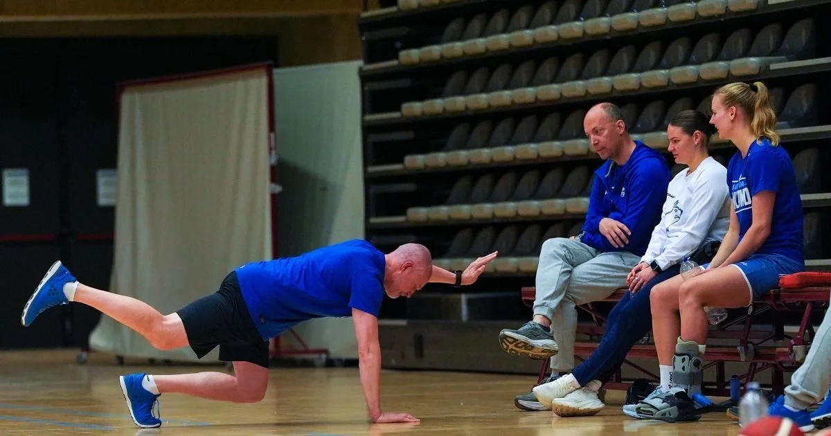 Henkilö tekee tasapainoharjoitusta urheilusalissa, muiden seuratessa suoritusta penkiltä.