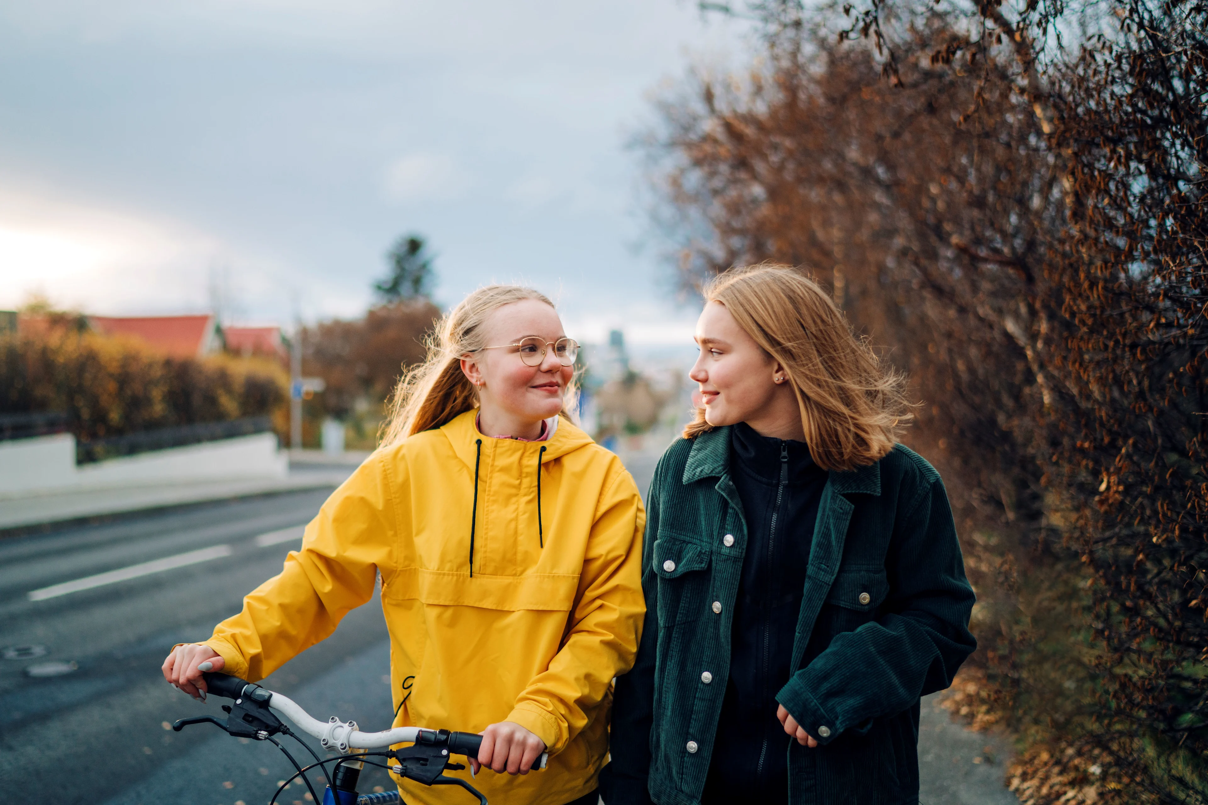 Kaksi nuorta kävelee syksyisellä kadulla vierekkäin ja hymyilee toisilleen, toinen pukeutuneena keltaiseen sadetakkiin ja toinen vihreään takkiin.