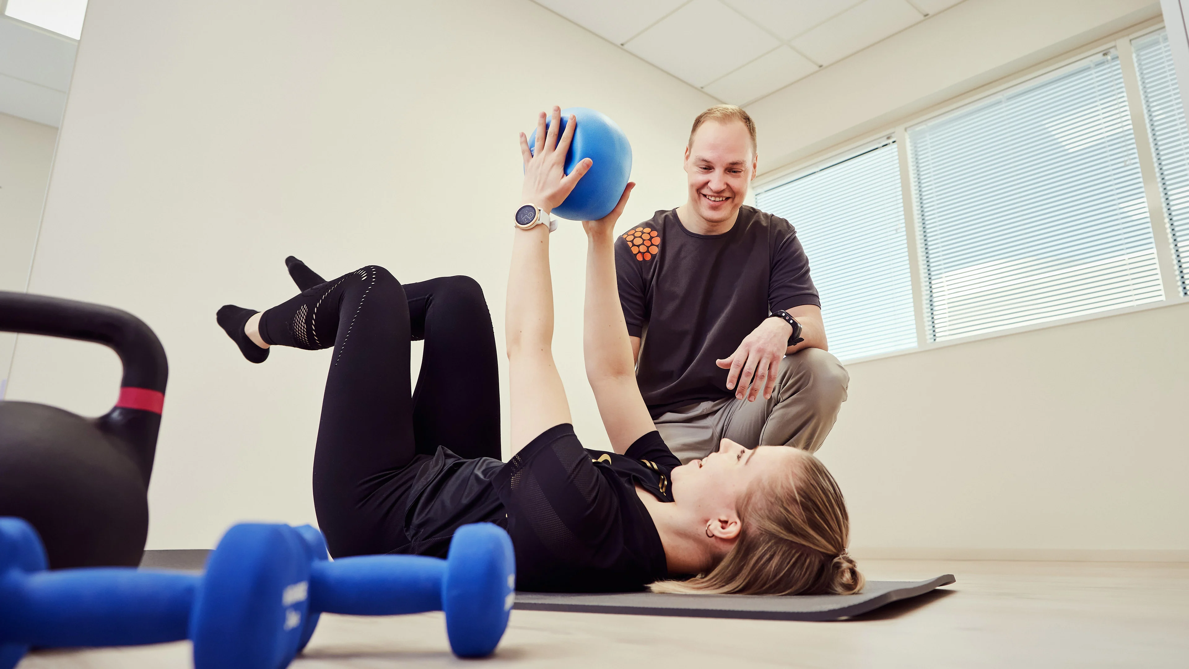 Työfysioterapeutti ohjaa asiakasta harjoituksessa jumppamatolla, välineinä kahvakuula ja käsipainot.