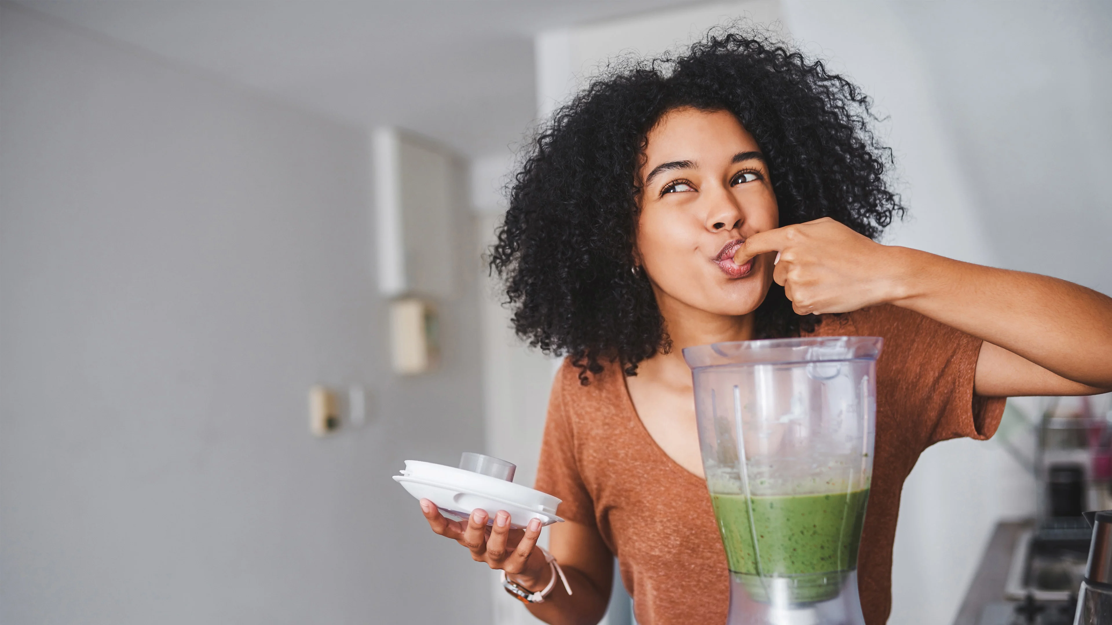 Hymyilevä nainen maistelee vihreää smoothieta sormestaan keittiössä. 