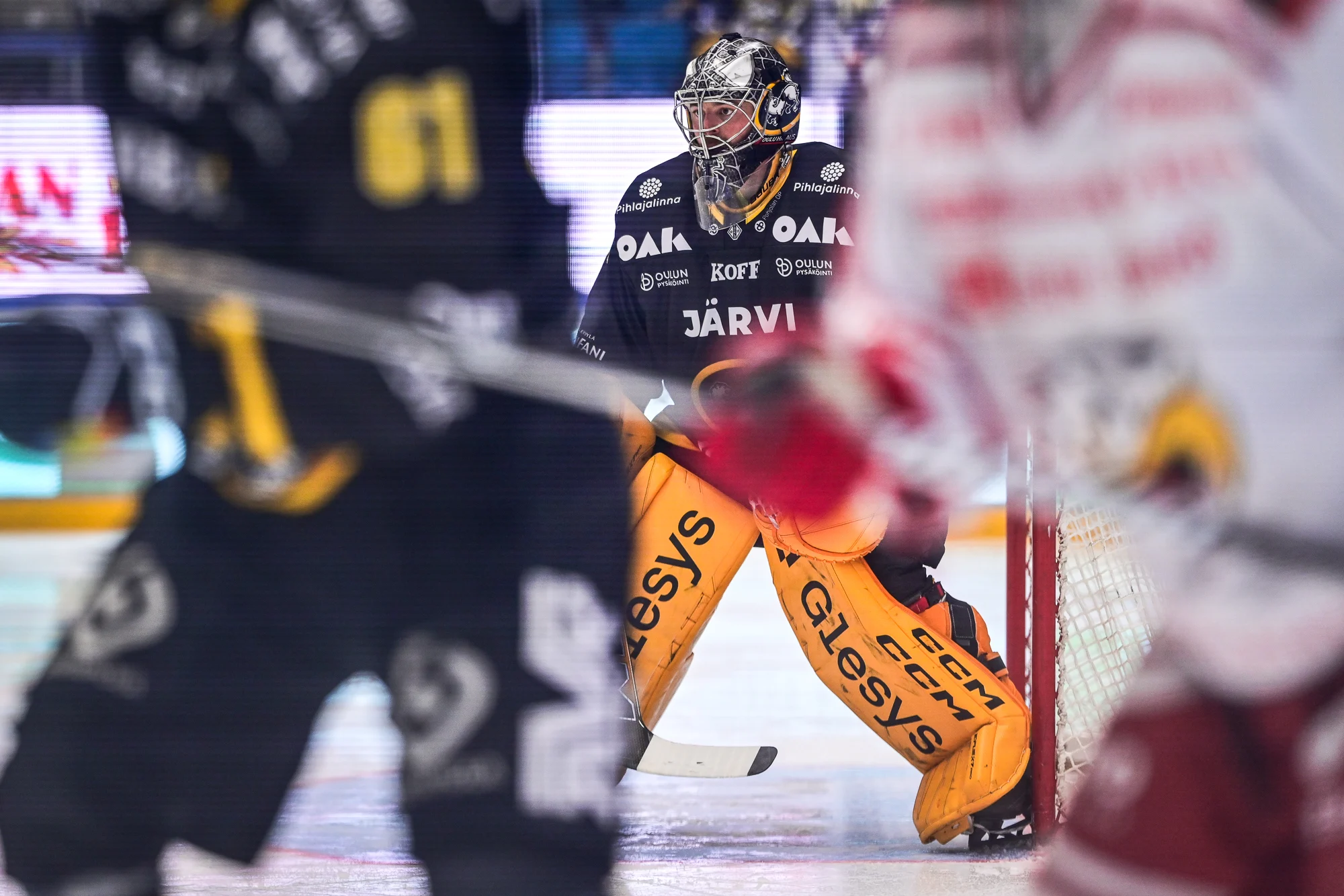 Oulun Kärppien maalivahti keskittyy työhönsä maalilla. Kuvan etualalla kaksi jääkiekon pelaajaa.
