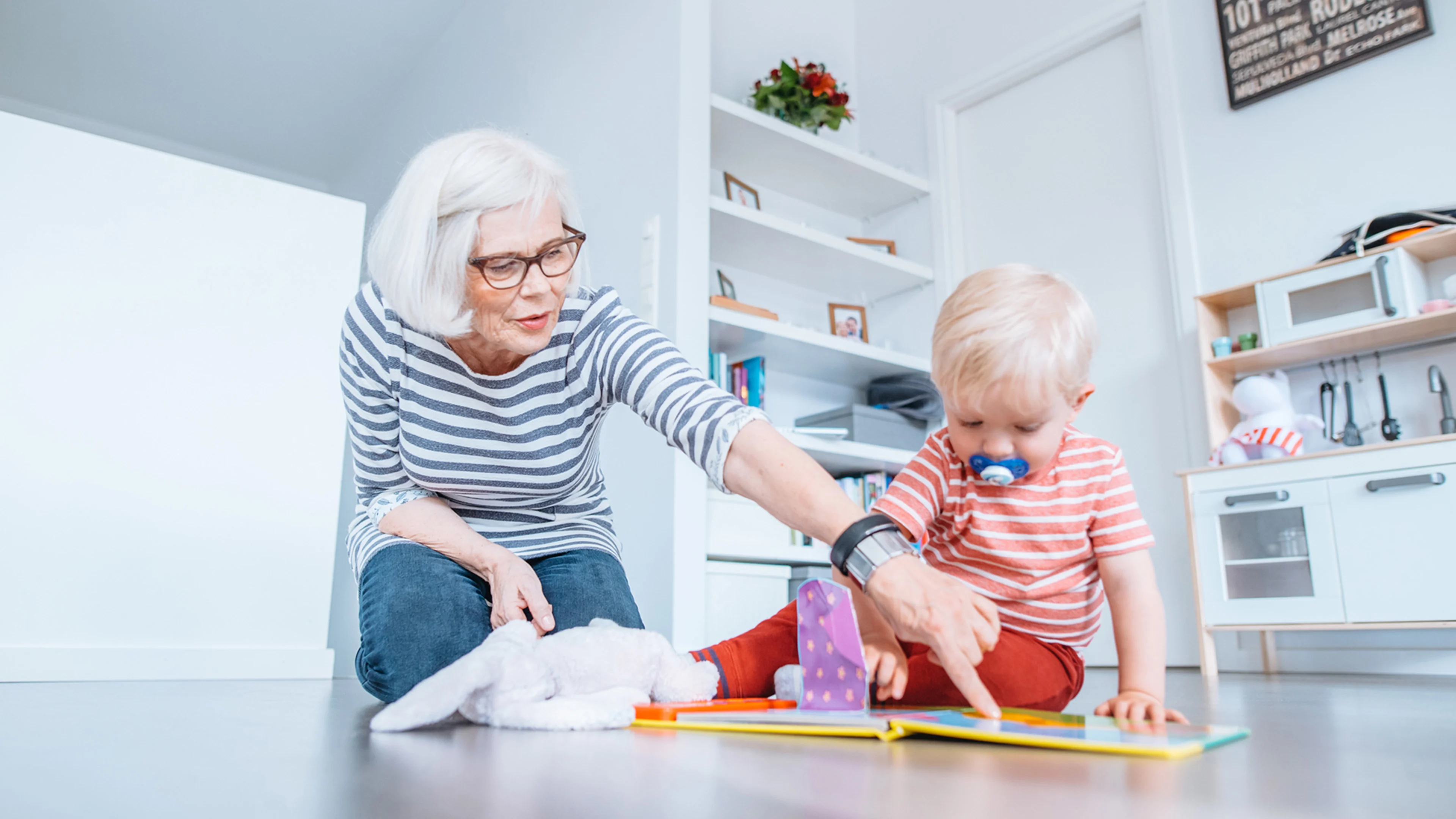 60-70 vuotias nainen ja noin 1 vuotias lapsi istuvat lattialla ja lukevat kirjaa. 