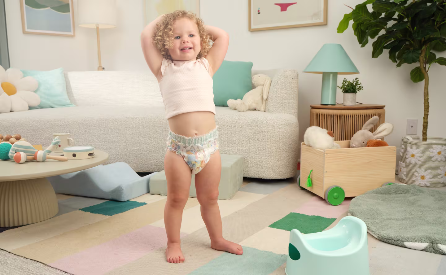 A smiling toddler with blonde curly hair standing in a colorful bright living room wearing her Easy Ups training pant. A couch, artwork, lamp and potty in the background