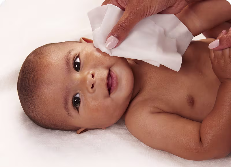 Close up of a laying baby smiling as mom gently wipes their face with a Pampers baby wipe Close up of a laying baby smiling as mom gently wipes their face with a Pampers baby wipe