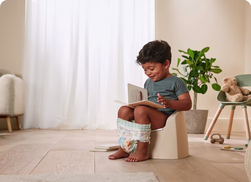 A toddler sitting on a potty reading a book, wearing Easy Ups A toddler sitting on a potty reading a book, wearing Easy Ups