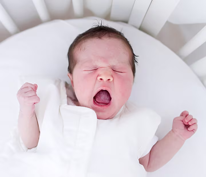 Yawning newborn baby lying in a white crib.