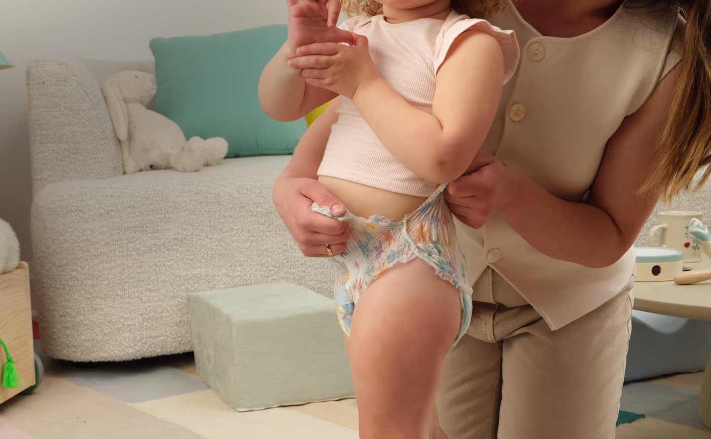 Close up of a toddler wearing Easy Ups, standing in a living room with mom kneeling beside her, carefully tearing the sides of her Easy Ups training pant. A coffee table with some wooden toys in the background