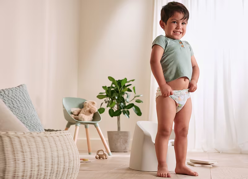 A smiling toddler wearing an Easy Ups training pant and green shirt stands in a well lit living room A smiling toddler wearing an Easy Ups training pant and green shirt stands in a well lit living room