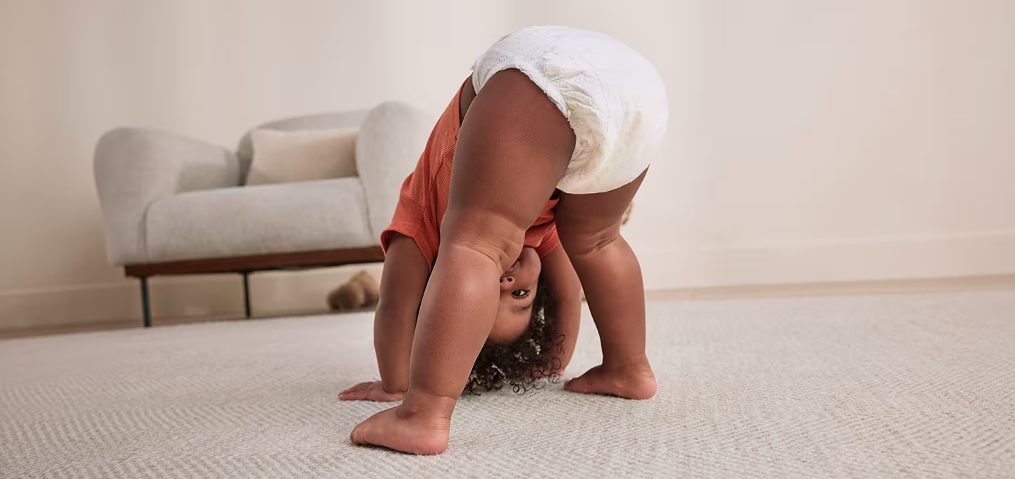 Toddler in diaper bends forward on carpet, peeking upside down between legs in a bright room.