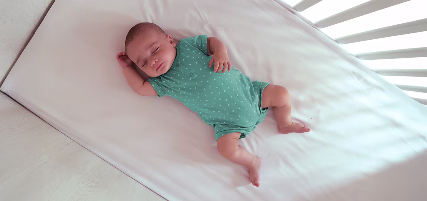 Baby calmly sleeping in a bare baby crib, wearing a teal pyjama with white dots
