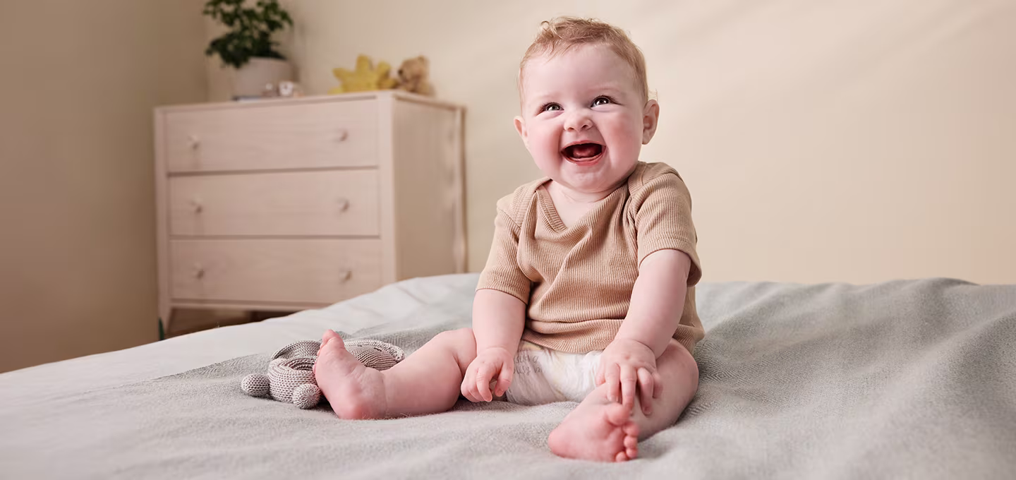 Smiling baby sitting on a bed holding a soft toy, showing a calm, self-soothing posture.