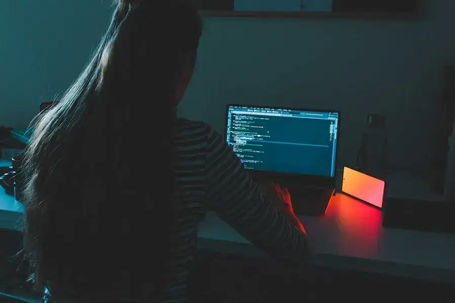a woman programming at her desk during the night time with the light of her computer and mobile phone 