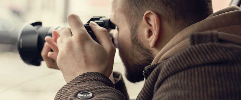 This is a photo of a male in a brown jacket. The man is taking a photograph. He appears to be spying on someone or trying to take the photograph discreetly. 