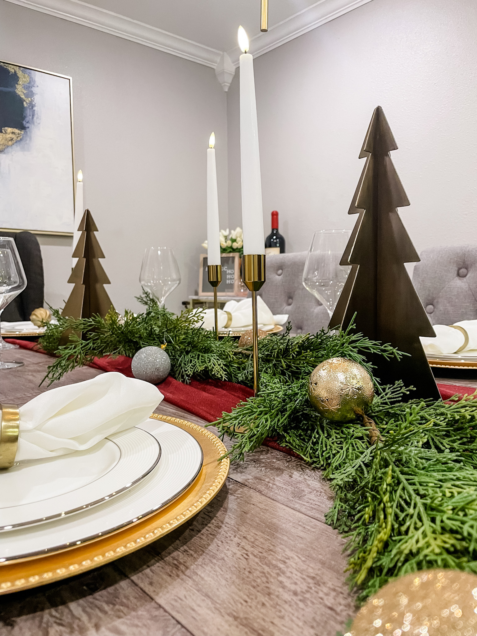 Holiday tablescape with white plates, maroon table runner, cedar pine garland, gold holiday trees, and flameless candles. 