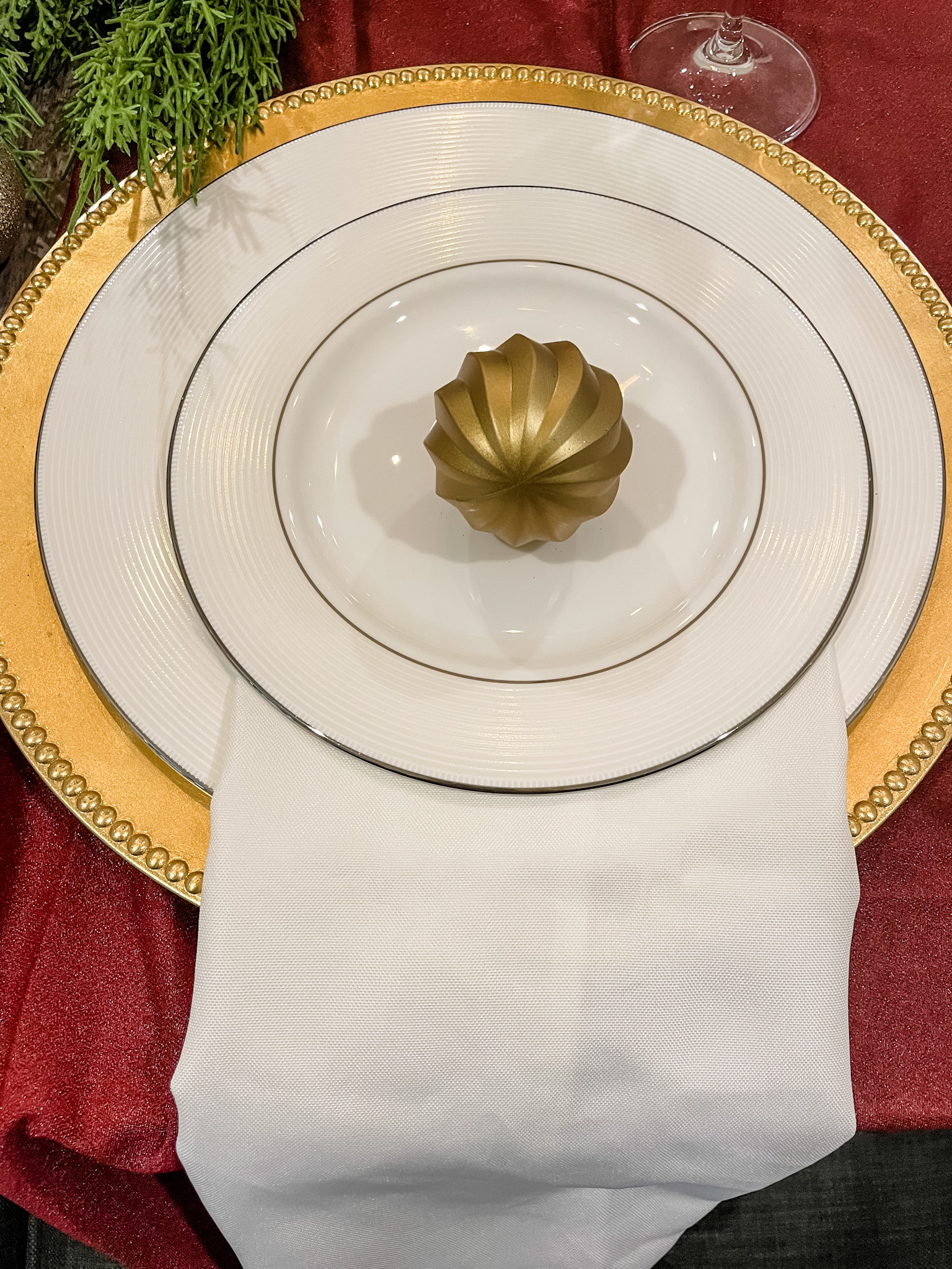 White china, with a gold charger, on a maroon table runner with a gold ornament on the plate. 