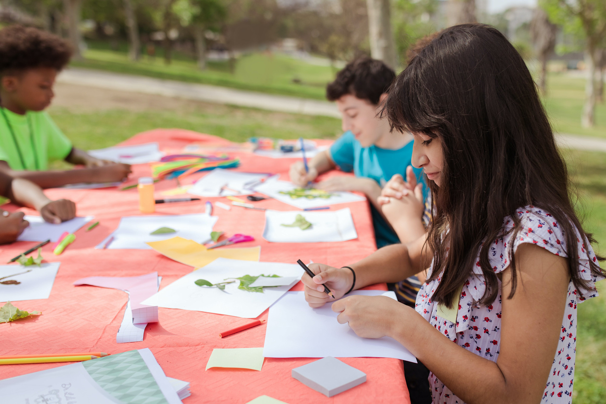 A group of young boys and girls sitting at a table outdoors, doing crafts. 