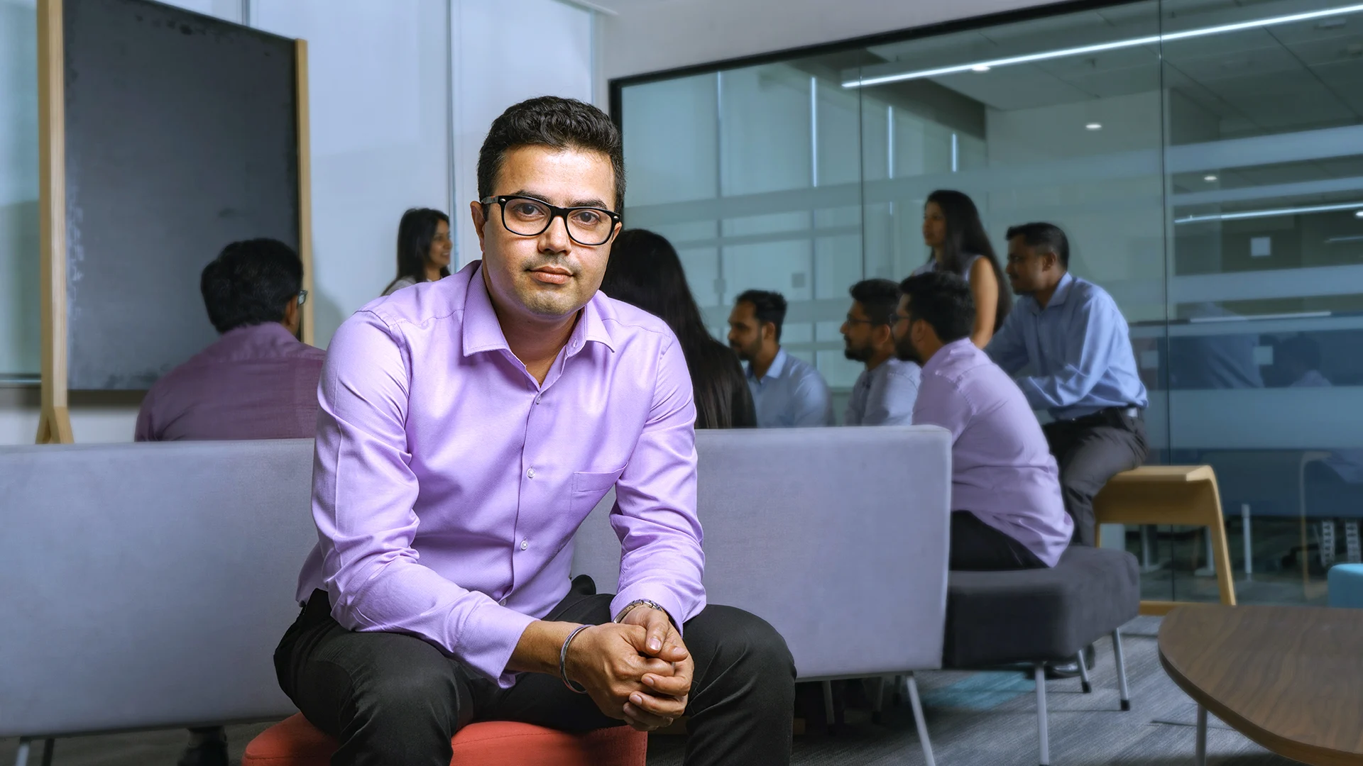 A male CSG employee sits in the Bangalore office as colleagues gather for discussion in the background.