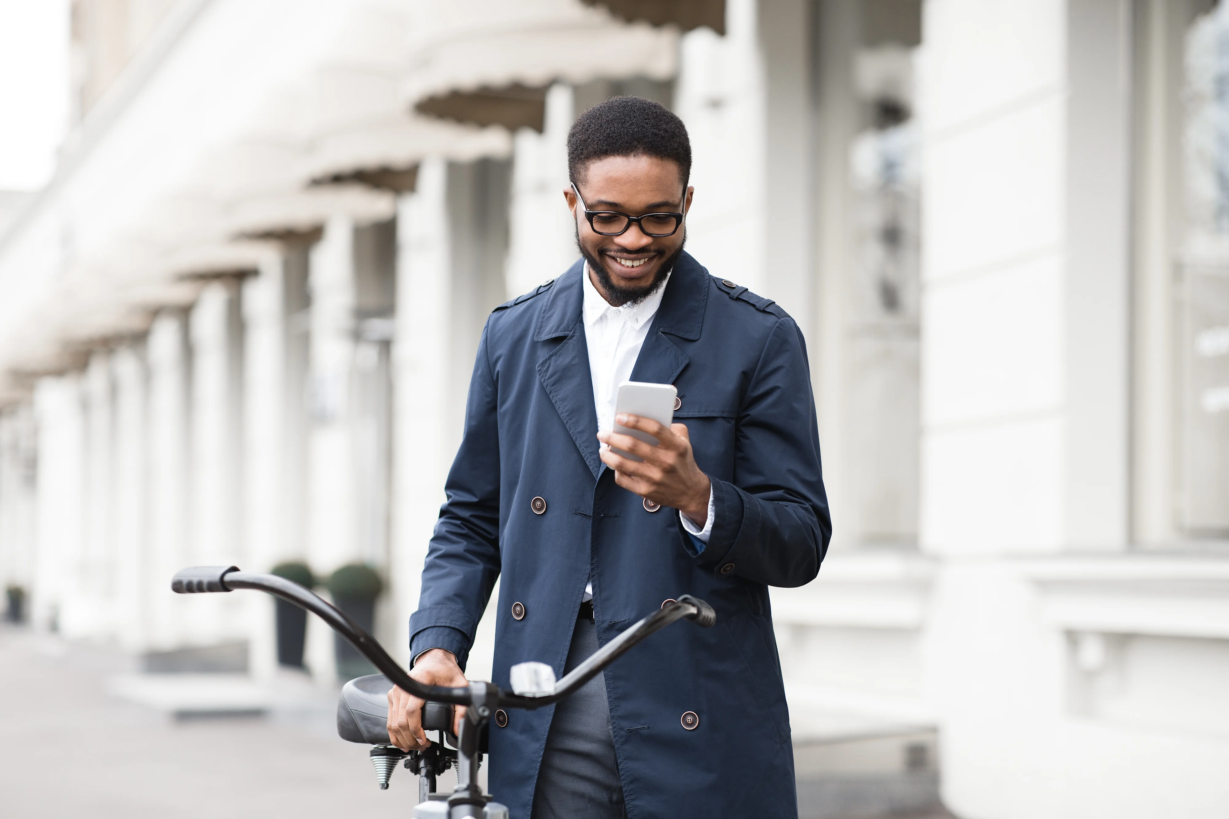 Young Black man texting on phone and standing with bike on city street.