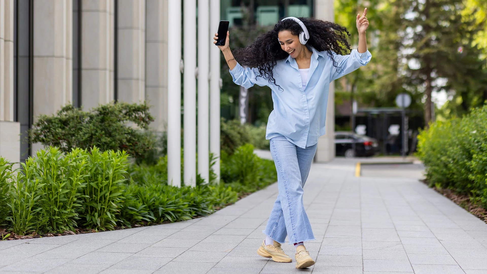 Young joyful beautiful woman dancing and singing in headphones while walking in the city