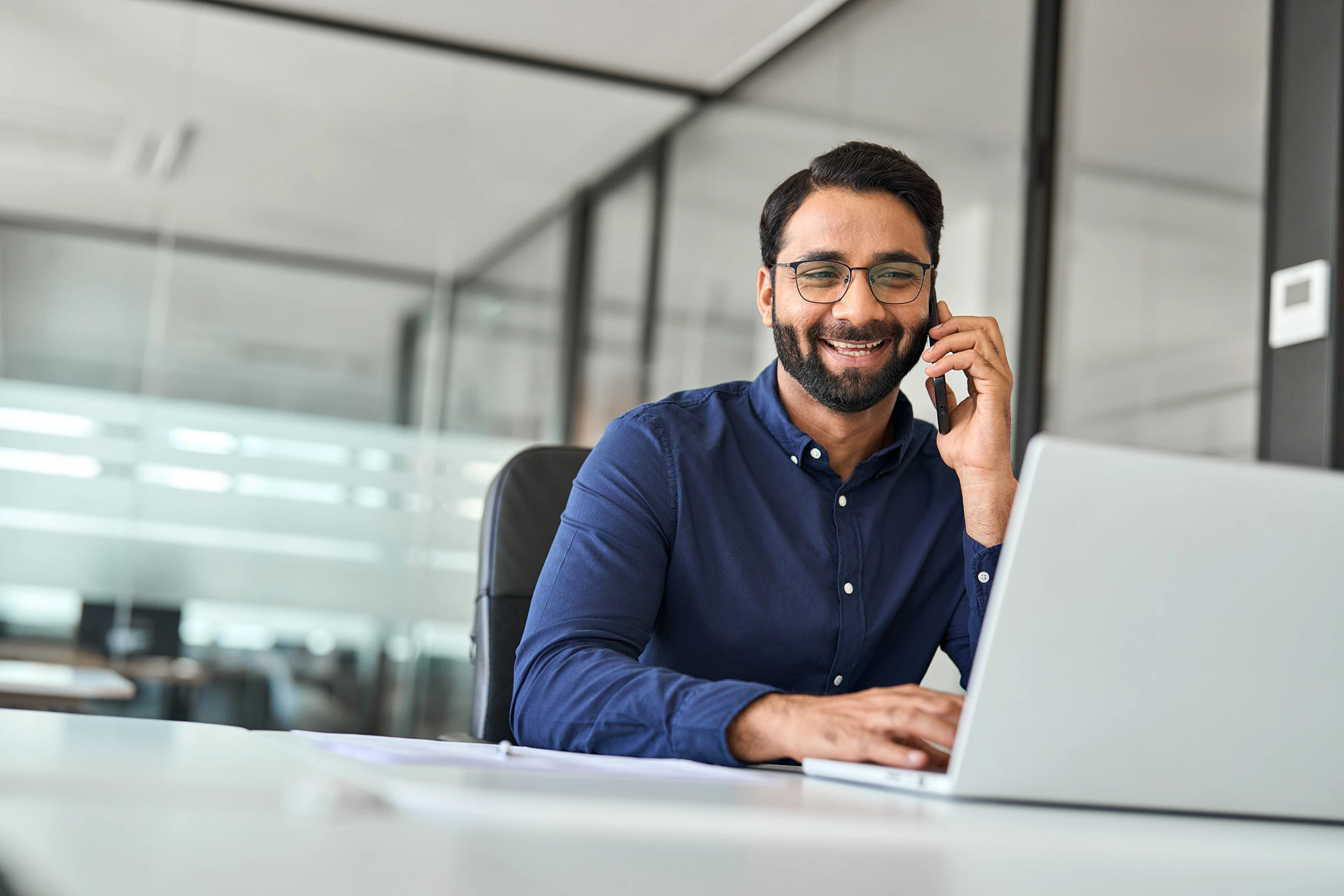 Happy Indian businessman merchant calling on cell phone using laptop in office.