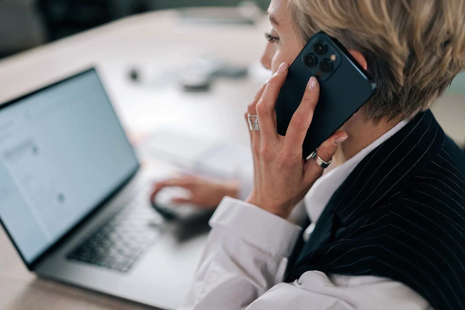 Closeup view from shoulder of business woman talking on mobile phone at workplace table, sitting at laptop computer, discussing work, making call from workplace.