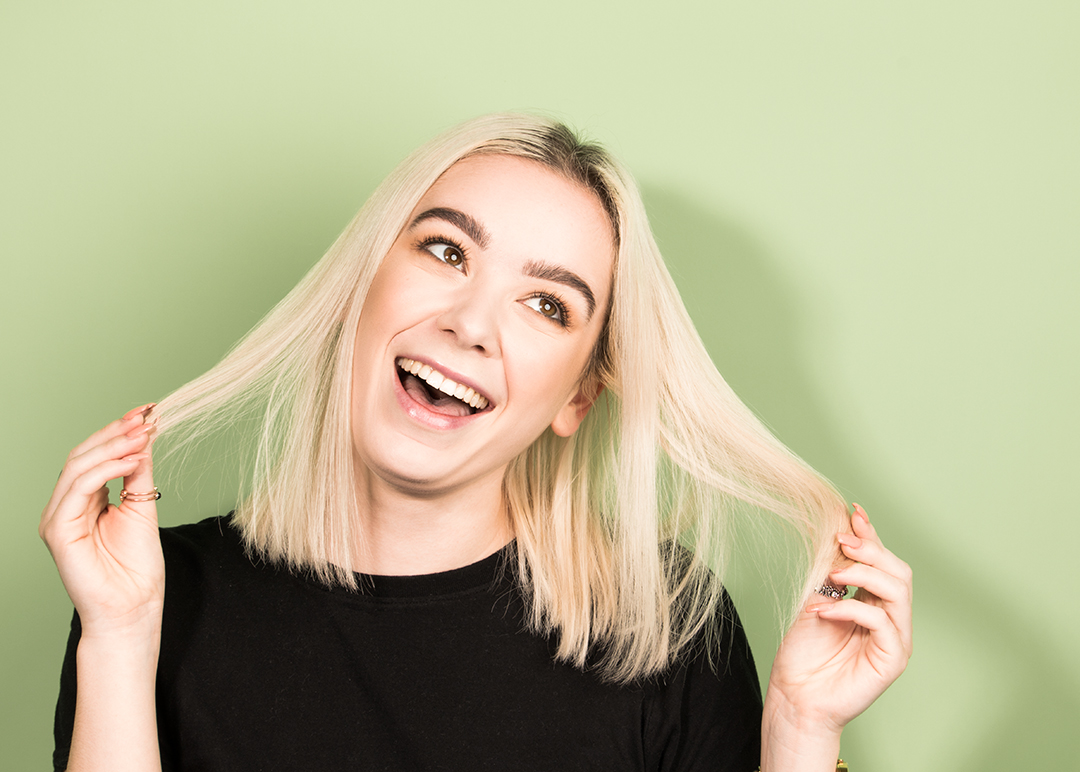 How to Dye Your Hair at Home Like a Professional - Young woman in front of light green background. She has with bleached blonde hair with dark roots and is holding out strands of her hair - 1080 x 772