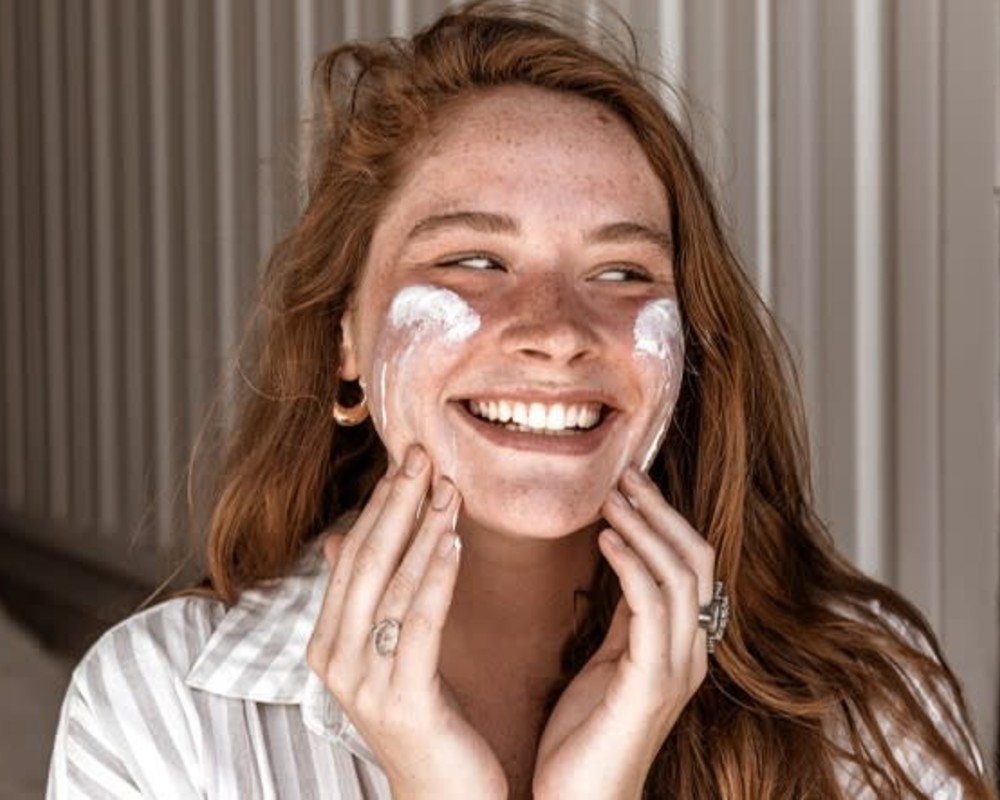 Ultimate Guide to Sunscreen - Woman with red hair and freckles smiles as she applies sunscreen to face