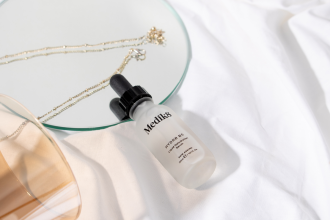 Adore Beauty - Medik8 Hydr8 B5 Liquid Rehydration Serum - glass product bottle with dropper lid lays flat on white sheets, small round mirror and a gold necklace  1200 x 800