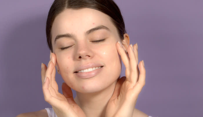 Best Skincare Routine for Oily Acne Prone Skin - young woman with her fingers on either side of her face had eyes closed and a few small pimples and a light purple background - 700 x 406