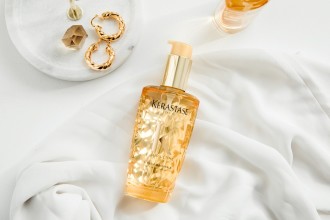 Kérastase Elixir Ultime Beautifying Hair Oil - product bottle flay lay on white material, next to a small white dish with gold jewellery - 1080 x 720
