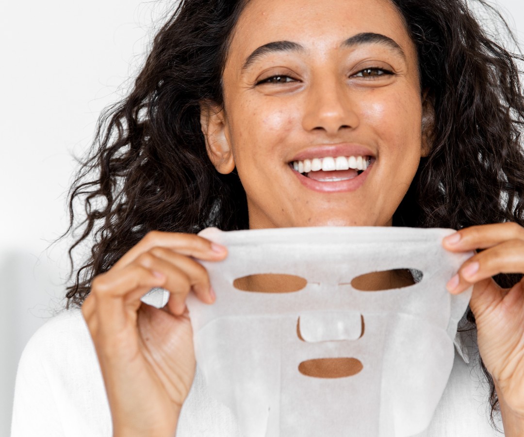 Best Face Masks for Dry Skin_woman smiling as she holds up a sheet face mask_1080x900
