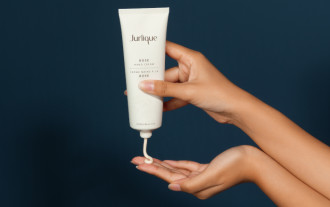 Jurlique Rose Hand Cream - one hand holds bottle while squeezing product into palm of other hand with a dark blue background- 700 x 440
