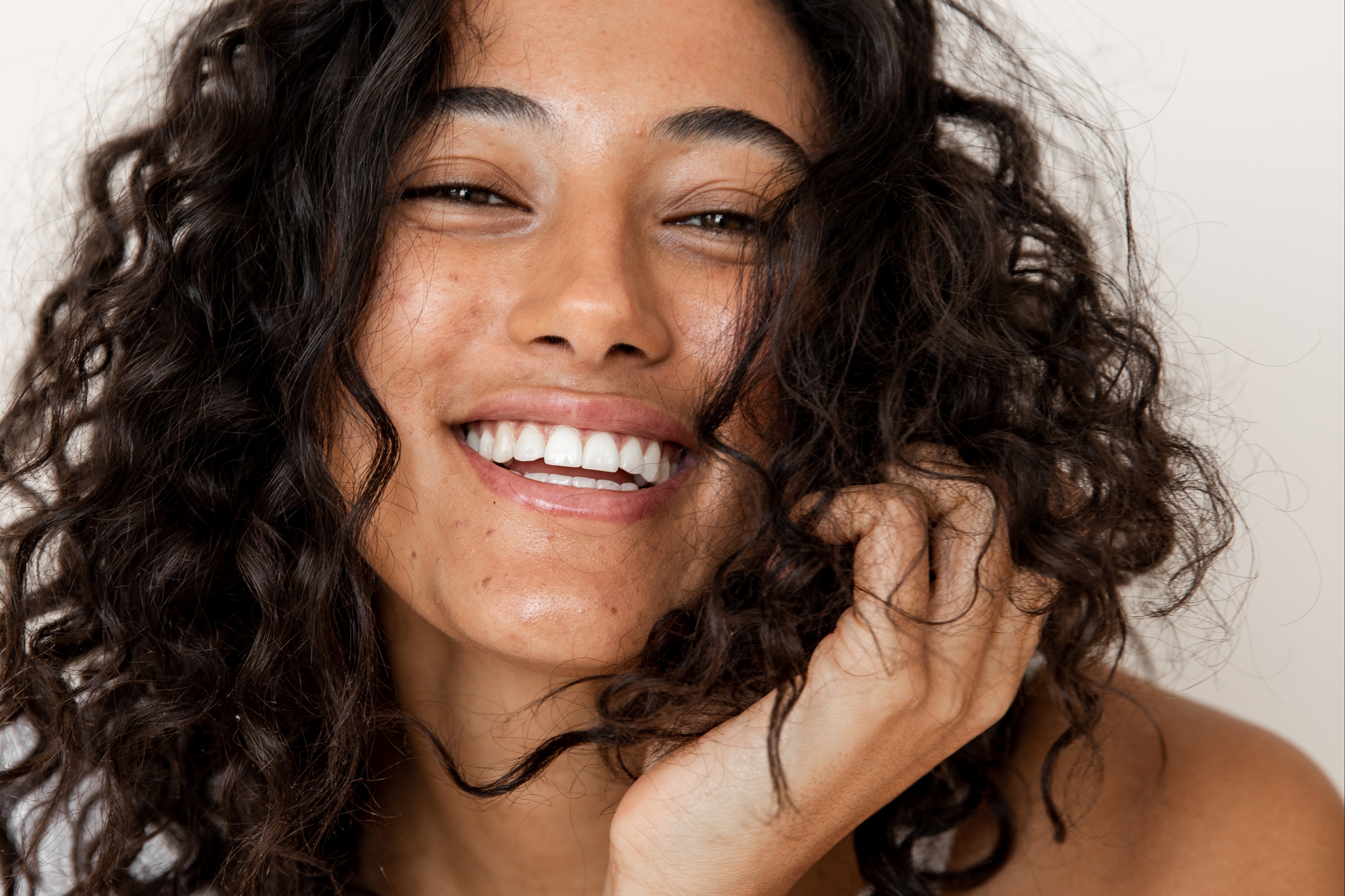 curly hair & frizz control - young woman with dark curly hair is smiling as her hand plays with the ends of her hair - 1200 x 800